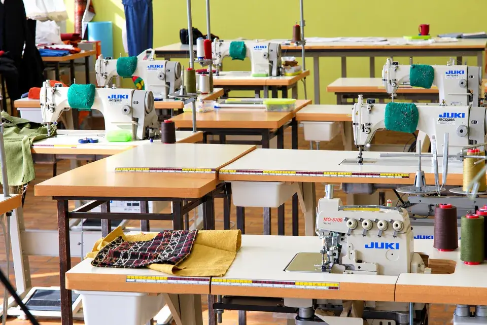 Dans l’atelier, une couturière assise entre deux rangées de machines à coudre industrielles ajuste un vêtement à carreaux mauves.