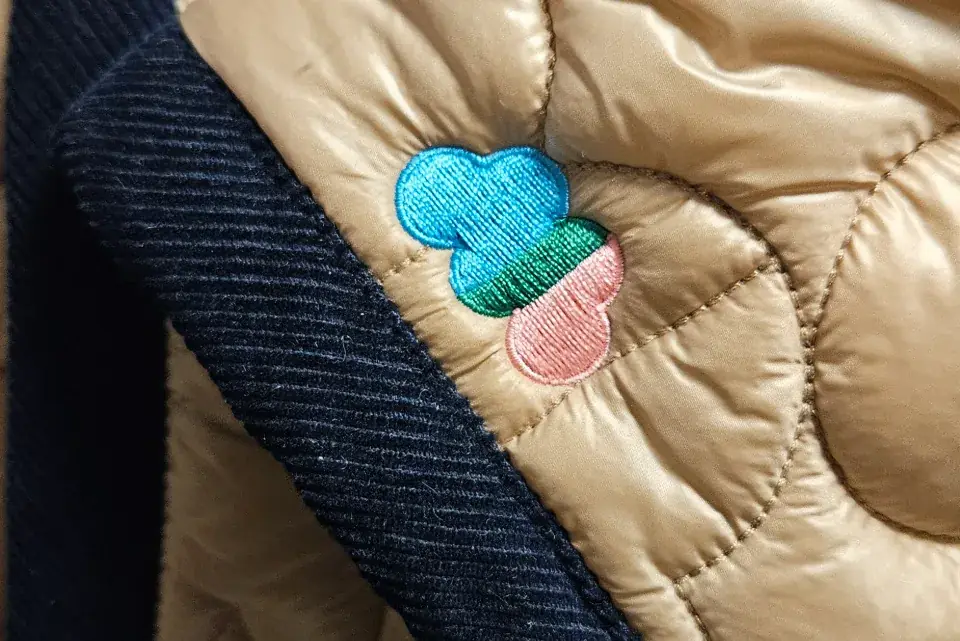 Une couturière souriante, mètre ruban autour du cou, présente une pièce de tissu rouge derrière sa machine à coudre industrielle.