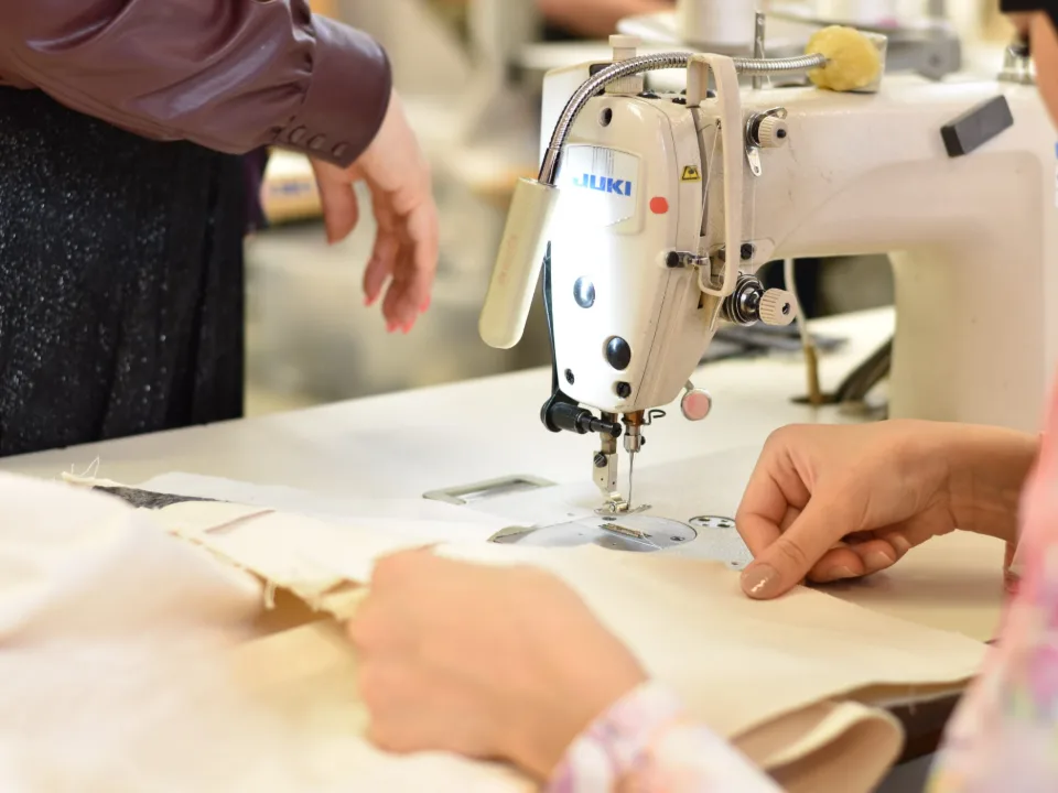 Deux mains, manches de veste de travail bleue, règlent au tournevis le pied presseur d’une machine à coudre industrielle.