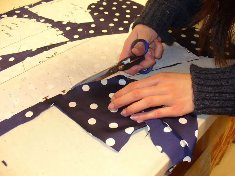 Découpe aux ciseaux d’un tissu bleu marine à pois blancs, épinglé sur son patron en papier pointé.