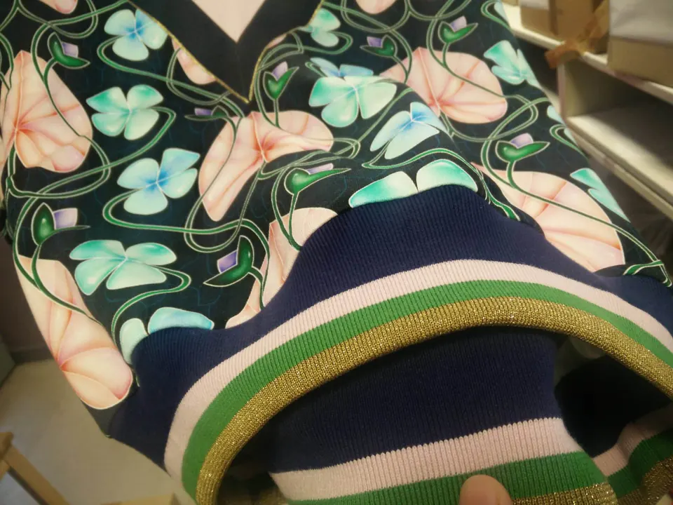 Détail d’un sweat imprimé de fleurs roses et turquoise, avec un bord-côte rayé vert, rose et fil doré.