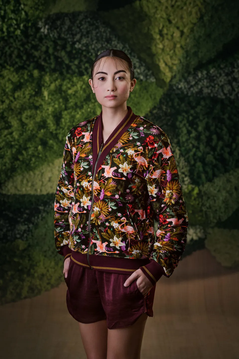Mannequin portant un blouson imprimé de fleurs tropicales et un short bordeaux devant un mur végétal.