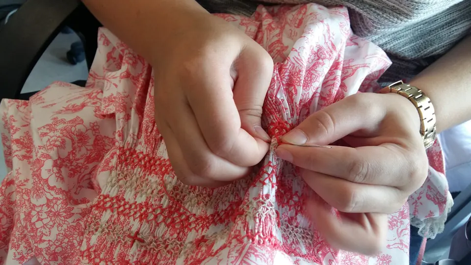 Des mains brodent à l’aiguille des smocks sur un tissu toile de Jouy rouge et blanc.
