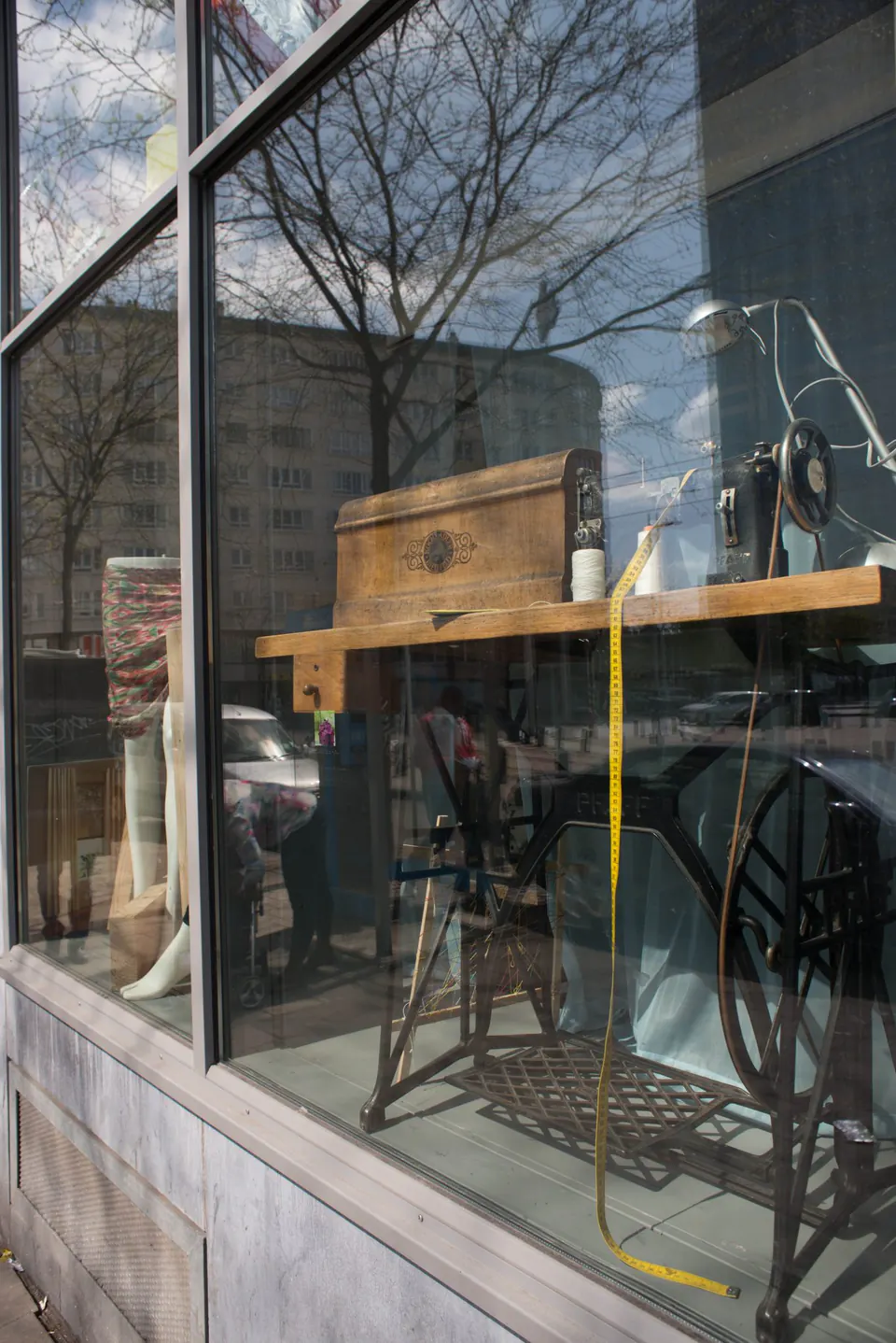 Etalage met een oude trapnaaimachine en een gele lintmeter; de straat en bomen weerspiegelen zich in het glas.